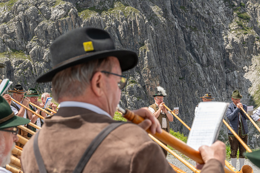 Male and female alphornplayers in traditional austrian clothing playing at the Lünersee mountain lake in Austria. Mountain slope in the background. Close up backview of a player. 8 july 2023, Brand, Austria