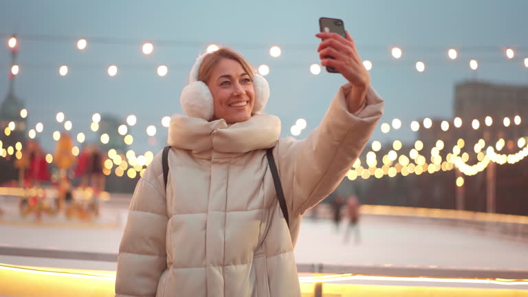 Happy woman talking selfie with smartphone outside ice rink during Christmas