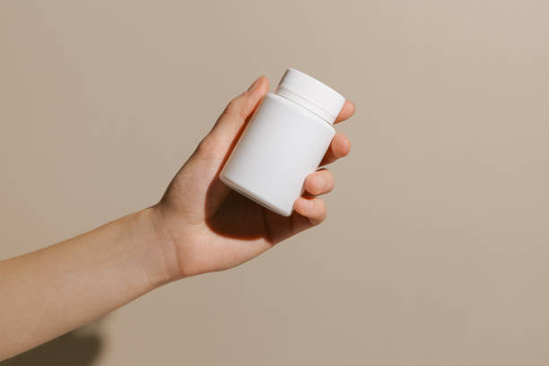 istock Female hand holding white small pill jar mockup on beige isolated background. Concept of pharmacy, health care, medicine, treatment. Image for your design. 2149383893