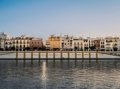 Colorful buildings including homes and shops face the Guadalquivir River in the Triana District of the Analusian city of Seville, Spain during golden hour summer sunset