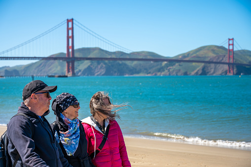 Two mature women and one man walking on beach of Presidio San Francisco and looking at view during springtime day, with Golden Gate bridge in background