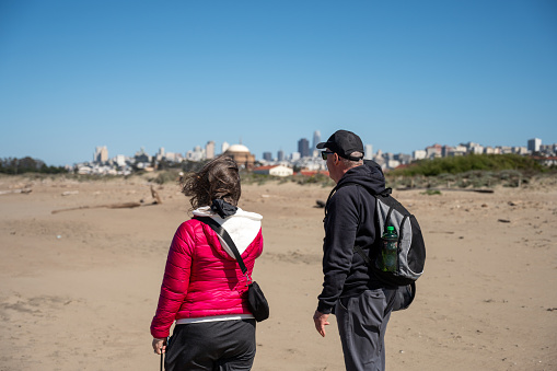Two mature man and woman walking on beach of Presidio San Francisco and looking at Skyline during springtime day