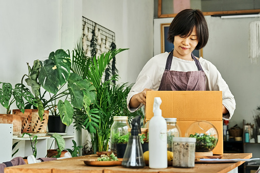 Asian woman packing terrariums in cardboard box for delivery while working at small garden center