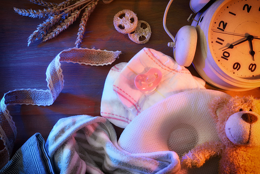 Conceptual background of bedtime for the baby with accessories on a table illuminated by a window at night on one side and with artificial light from the room, accessories for the baby and alarm clock on the other.