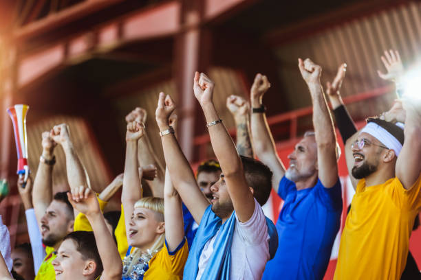 Closeup of hands in the air. Sport fans cheering at the game on stadium. Wearing yellow and blue colors to support their team. Celebrating with flags and scarfs.