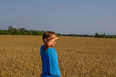 In a wheat field on a summer day, a child looks into the distance, covering his eyes with his hand. Shielding his eyes from the bright summer sun, the child looks at the wheat field.