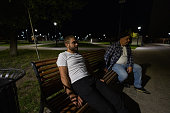 Two pensive men sit on a bench in the park at night in the summer. Two friends are resting thoughtfully on a summer evening in a city park. Men in the park are sitting on a bench.