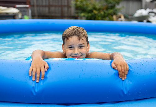A handsome boy smiles sweetly at the camera while swimming in an inflatable pool. The child cutely puts his head on the edge of the pool and smiles while looking at the camera..