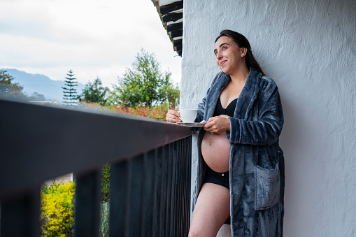 Beautiful pregnant Latin woman from Bogotá Colombia between 20 and 29 years old, enjoys drinking her coffee next to the window while wearing her nightgown