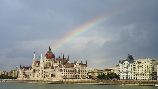 A natural arch of colors crowns the Hungarian Parliament Building in Budapest, a marvel of architecture. The majestic structure sits by the Danube River, with the rainbow's ephemeral beauty adding a touch of magic to the historic skyline.