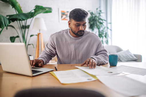 Focused indian man at home using a laptop with documents on desk and coffee mug alongside