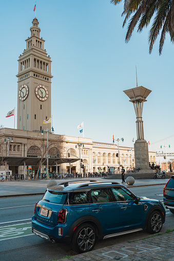 San Francisco, California, April 8, 2024. A blue compact car parked on the street with the Ferry Building clock tower rising in the background.