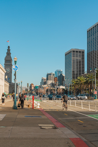 San Francisco, California, April 8, 2024. Urban street scene at The Embarcadero with cyclists and pedestrians, flanked by high-rise buildings and palm trees.