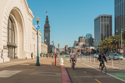 San Francisco, California, April 8, 2024. Urban street scene at The Embarcadero with cyclists and pedestrians, flanked by high-rise buildings and palm trees.