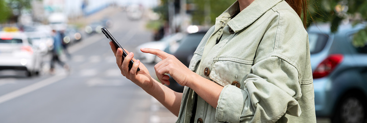 Female urban person stands next to road in city and using her smartphone. Searching car rental, taxi, public transportation.