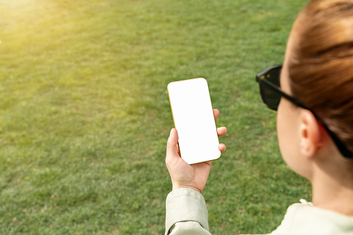 Woman holding smart phone with blank white screen in her hand against green lawn. Mockup screen of phone over the shoulder.