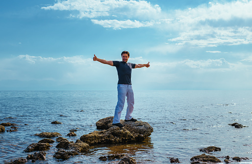 Handsome successful man standing on rock in the middle of lake and showing thumbs up