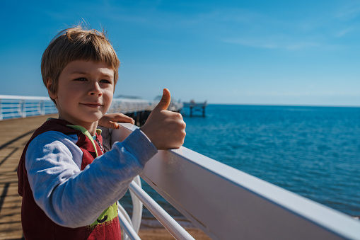 Smiling boy shows thumbs up standing on the pier