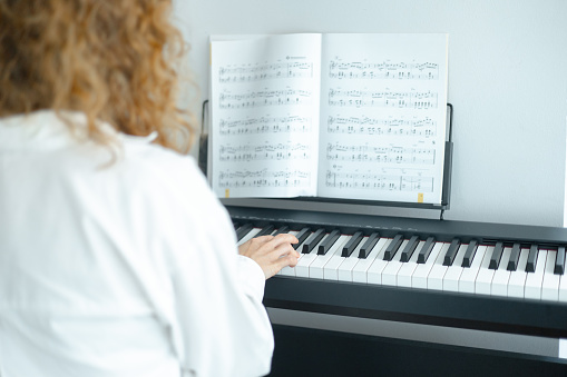 Young woman in white clothes sitting and playing an electronic piano with music notes. View from back