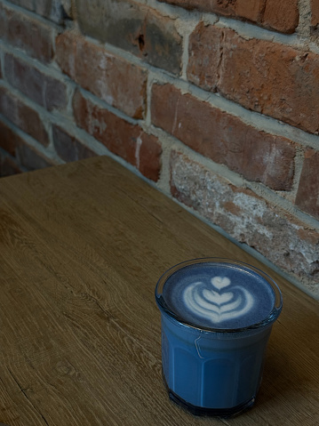 A refreshing blue drink with heartshaped latte art sits on a wooden table in front of a rustic brick wall, creating a cozy and charming atmosphere