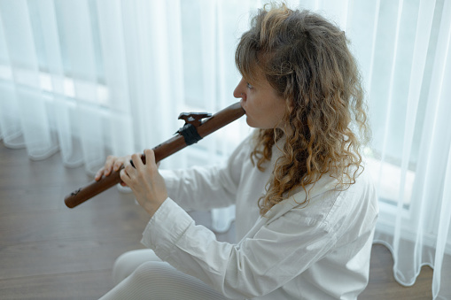 Beautiful young curly woman playing a wooden flute at home near the window, wearing in white outfit