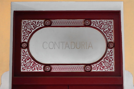 Cienfuegos, Cuba-October 11, 2019: Maroon-crimsonish painted Contaduria -Accountancy- door with a glazed upper transom window and wood filigree decoration, in the AD 1889 built Tomas Terry T.