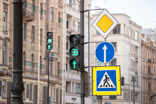 A traffic light on green with a timer, 'Main Road' and 'Straight Ahead' traffic signs, and a pedestrian crossing indicator against city buildings.