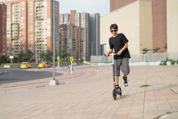 Teenager between 12 and 15 years old playing with his scooter next to a large concrete building enjoying a sunny day and the vitality of his body, He is wearing a summer outfit with shorts and a t-shirt. urban city lane.