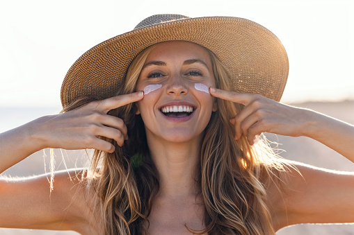 Portrait of beautiful smiling woman applying sunscreen on her face while looking at camera at the beach.