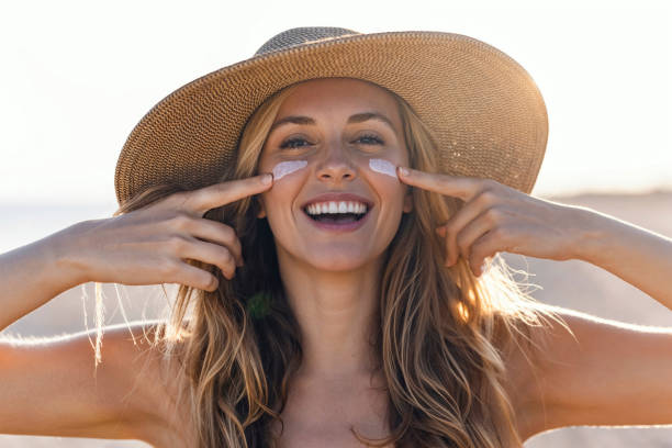 Beautiful smiling woman applying sunscreen on her face while looking at camera at the beach. Portrait of beautiful smiling woman applying sunscreen on her face while looking at camera at the beach. suntan lotion stock pictures, royalty-free photos & images