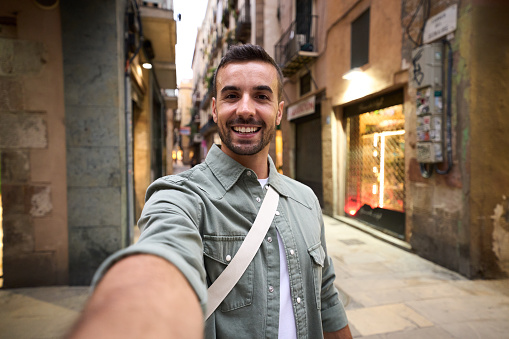 Young adult happy Caucasian man taking smiling selfie on Madrid touristic city street in background. Millennial handsome tourist takes lighthearted photo outdoors on his vacation trip in Europe