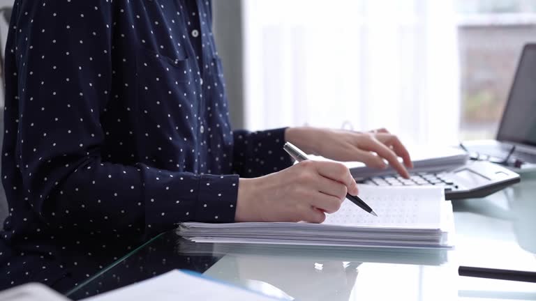 Businesswoman using calculator and reviewing ring folder of financial documents with magnifying glass at desk in modern office. Audit and taxes in business