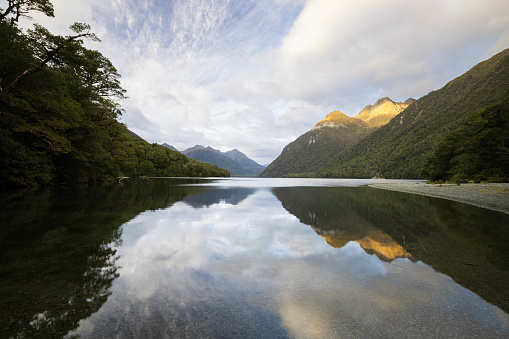 The early morning sun lights up the tip of the mountains surrounding Lake Gunn. The scene is reflected in the still waters.\nLake Gunn is situated in the Fiordland district of New Zealand's South Island. It is surrounded by mountains and native bush. Being situated just off the road into Milford Sound, it is often visited by tourists on their way to Milford Sound. The view here is from the north end of the lake.