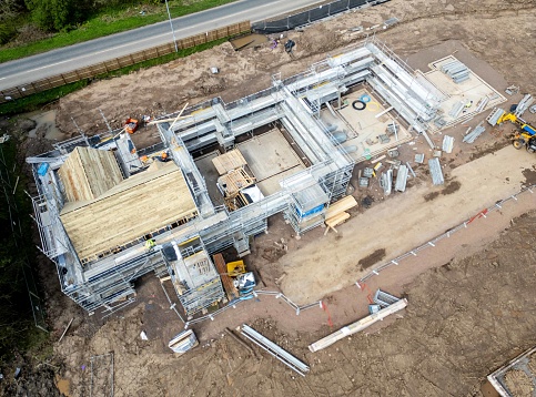 Overhead aerial drone view of new homes being built on a greenfield site in Midlothian, Scotland. Scaffolding surrounds the new house structures, as construction workers build up walls and put new roofs on, ready for tiles to be added.