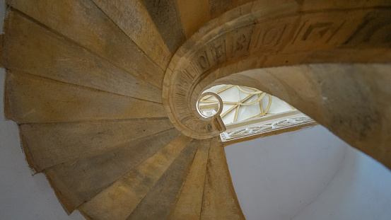 The graceful curve of a spiral staircase within a relocated church in Most, Czechia. Warm light bathes the stone steps, highlighting their texture and the intricate carvings that adorn them. The perspective offers a glimpse of architectural beauty, leading the eye upwards to the celestial window above.