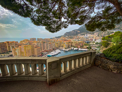 The lookout view is framed by a tree branch and the stone fence in the foreground. Buildings surround the marina. A mountain appears in the right background.
