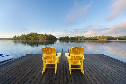 Two yellow Adirondack chairs on a wooden dock welcome the serene Muskoka summer morning, overlooking the blue waters of a lake. Across the way, cottages nestled among green trees complete the picturesque scene.