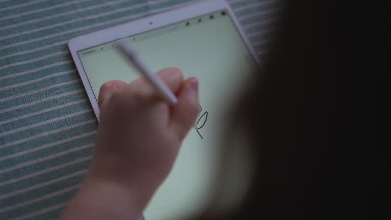 View from shoulder of unrecognizable child sitting at a table in front of modern digital graphics tablet with stylus in hand, drawing using technological device.