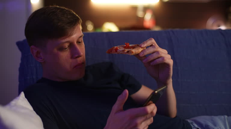 Handsome man eating pizza, browsing apps on smartphone, using online internet on mobile phone lying on comfy sofa at home in evening. Young male using mobile phone