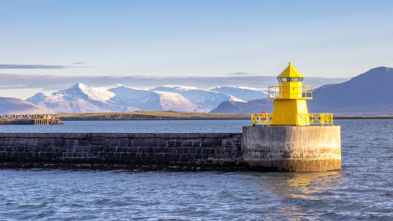 Panoramic view of a yellow lighthouse and harbour wall in Reykjavik at sunrise. Early morning with snow covered mountains in the distance. Iceland.