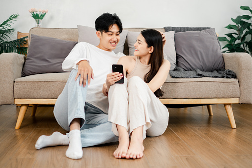 Young Asian couple sits cozily on a beige sofa, sharing a moment with a smartphone in a well-lit, modern living room.