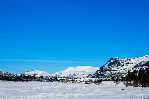 Winter in Stora Sjöfallet National park. The river is frozen and has tracks of snow mobiles. In the background the snow covered mountains. Blue sky.