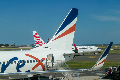 Planes of the four main domestic airlines in Australia at Sydney Kingsford-Smith Airport. In the foreground is a Rex Airlines Boeing B737-8FE, registration VH-RYU. Behind her is a Virgin Australia Boeing B737-8FE plane, registration VH-YVA. Between the two vertical stablisers is a Qantas Airbus A330-202 plane, registration VH-EBP.  In the left distance is a Jetstar Airbus A320-232 plane. This image was taken inside the domestic terminal on a sunny afternoon on 1 April 2024.
