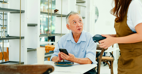 Elderly Asian male customer uses her smartphone to make a mobile payment at modern cafe, illustrating modern payment convenience. Small business owner concept.