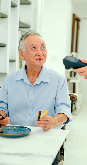 Elderly Asian male customer uses her smartphone to make a mobile payment at modern cafe, illustrating modern payment convenience. Small business owner concept. Vertical Screen.