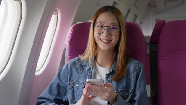Happy young woman sitting alone on her window seat on an airplane using her phone, looking up and smile at camera