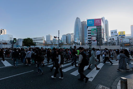 A large number of people, including tourists, cross the scramble crossing in front of the East Exit of Shinjuku Station in Tokyo on April 19, 2024. In the background, buildings in the foreground are being demolished to make way for the West Exit redevelopment project, and buildings that were not previously visible can be seen.