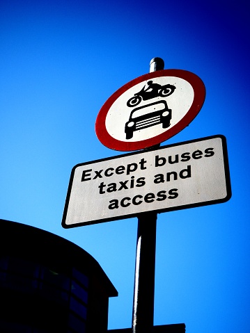 UK roadsign with car and motorbike symbols in Red circle above sign saying 'Except buses taxis and access'. Set against silhouetted building and deep blue sky