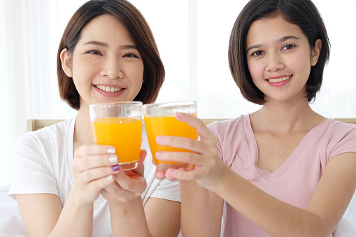 Smiling Asian young sisters lovely couple on white bed and smiling with hand holding Orange juice glass in bedroom. Holiday and healthy concept