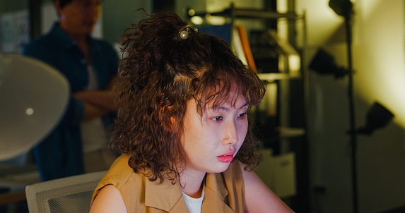 Young woman with curly hair intently focuses on her laptop screen, working late in a dimly lit office setting. ESG sustainable business office night concept.